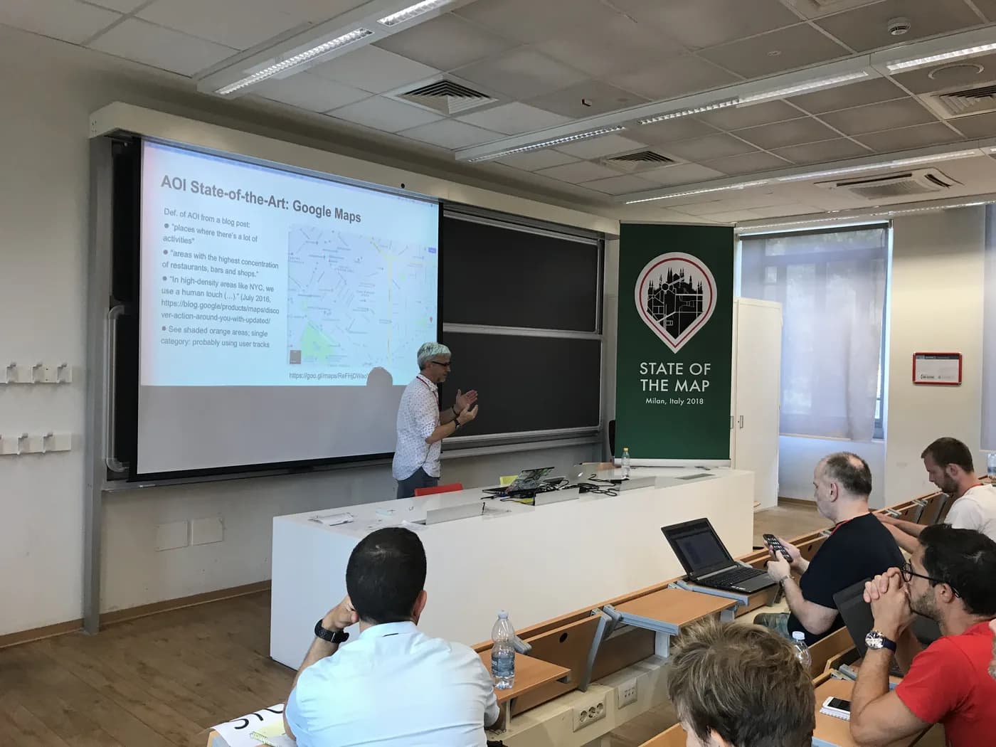 A speaker presenting a slide about Google Maps areas of interest beside a State of the Map Milan 2018 banner.