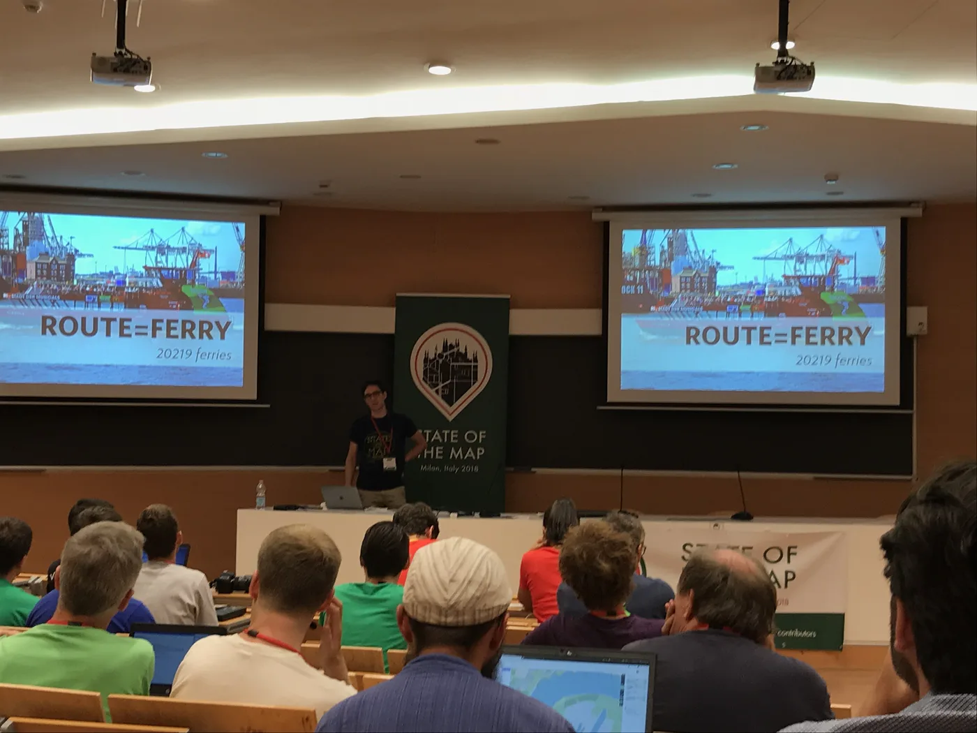 A State of the Map talk titled Route equals Ferry, shown on two projection screens.