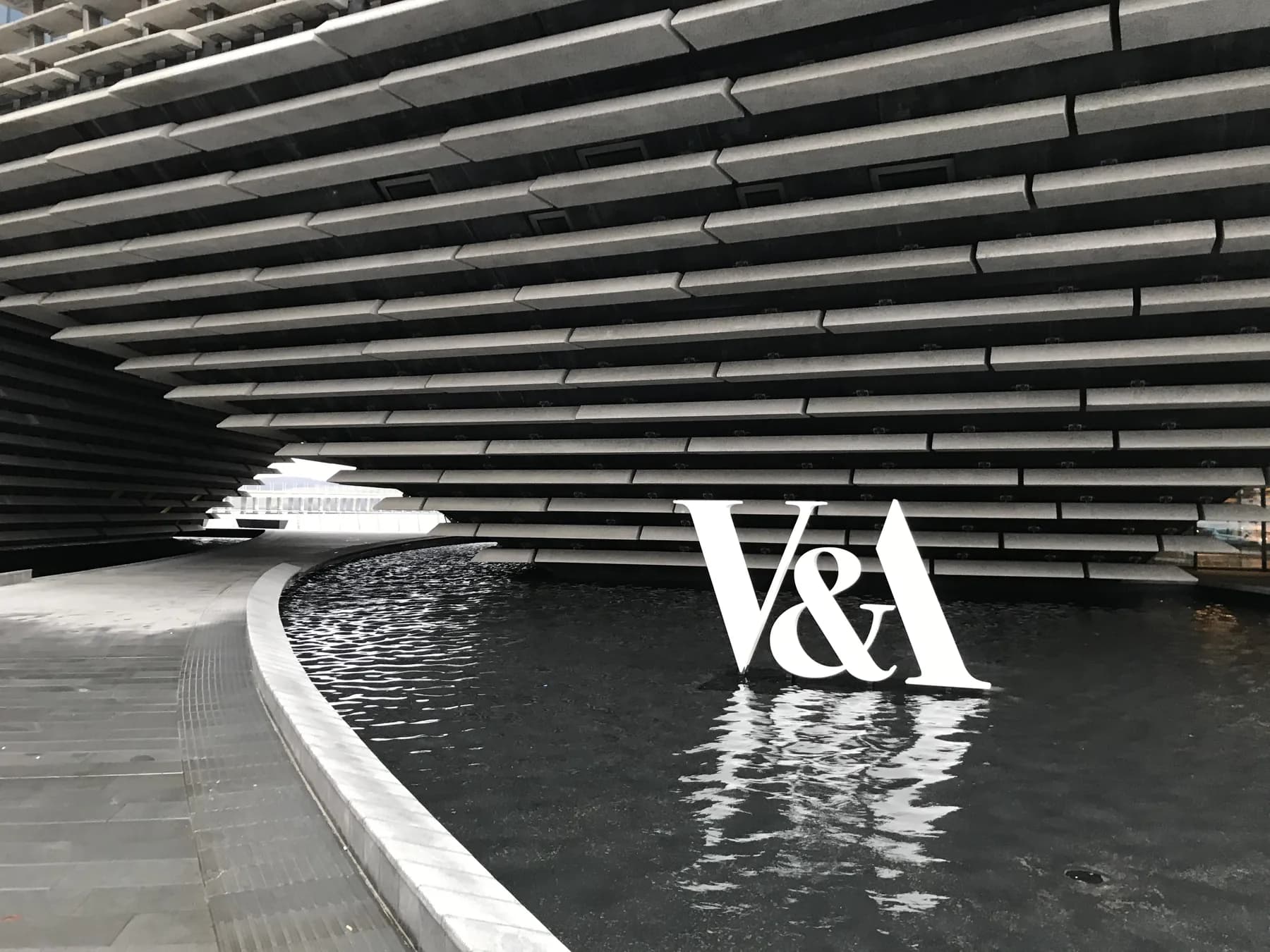 V&A Dundee's dark layered exterior above a reflecting pool, with a white V&A logo in the water.
